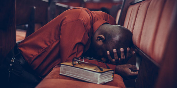 Homem ajoelhado no chão, apoiando-se no banco de uma igreja. Representa nossa lista de versículos sobre pecado.